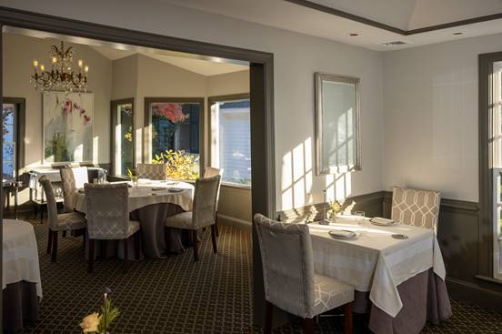 Elegant restaurant dining room with tables covered in white linens, cushioned chairs, soft natural light coming through large windows, and a chandelier hanging from the ceiling.