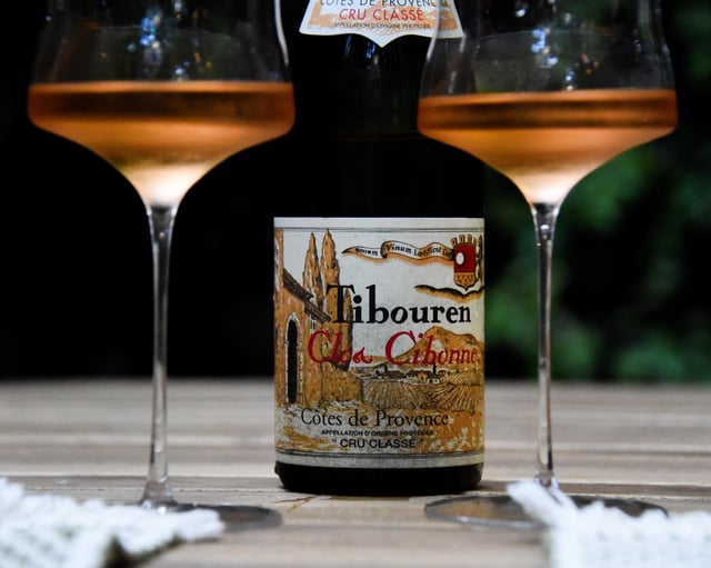 A bottle of Tibouren Clos Cibonne rosé wine sits on a wooden table between two filled wine glasses, with part of the wine label and two white napkins in view.