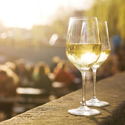 Two glasses of white wine rest on a stone ledge outdoors, with a sunlit, blurred background of people sitting and socializing in a garden or patio setting.