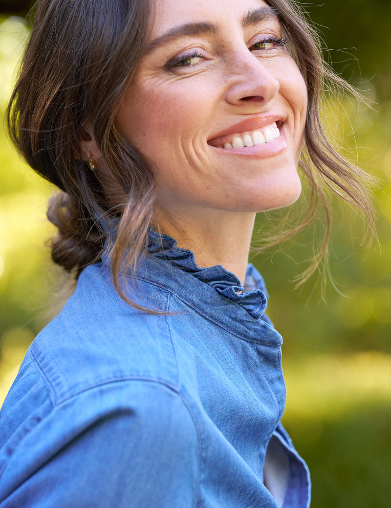 A woman with wavy brown hair in a low bun smiles warmly at the camera while wearing a blue denim shirt, outdoors with a blurred green background.