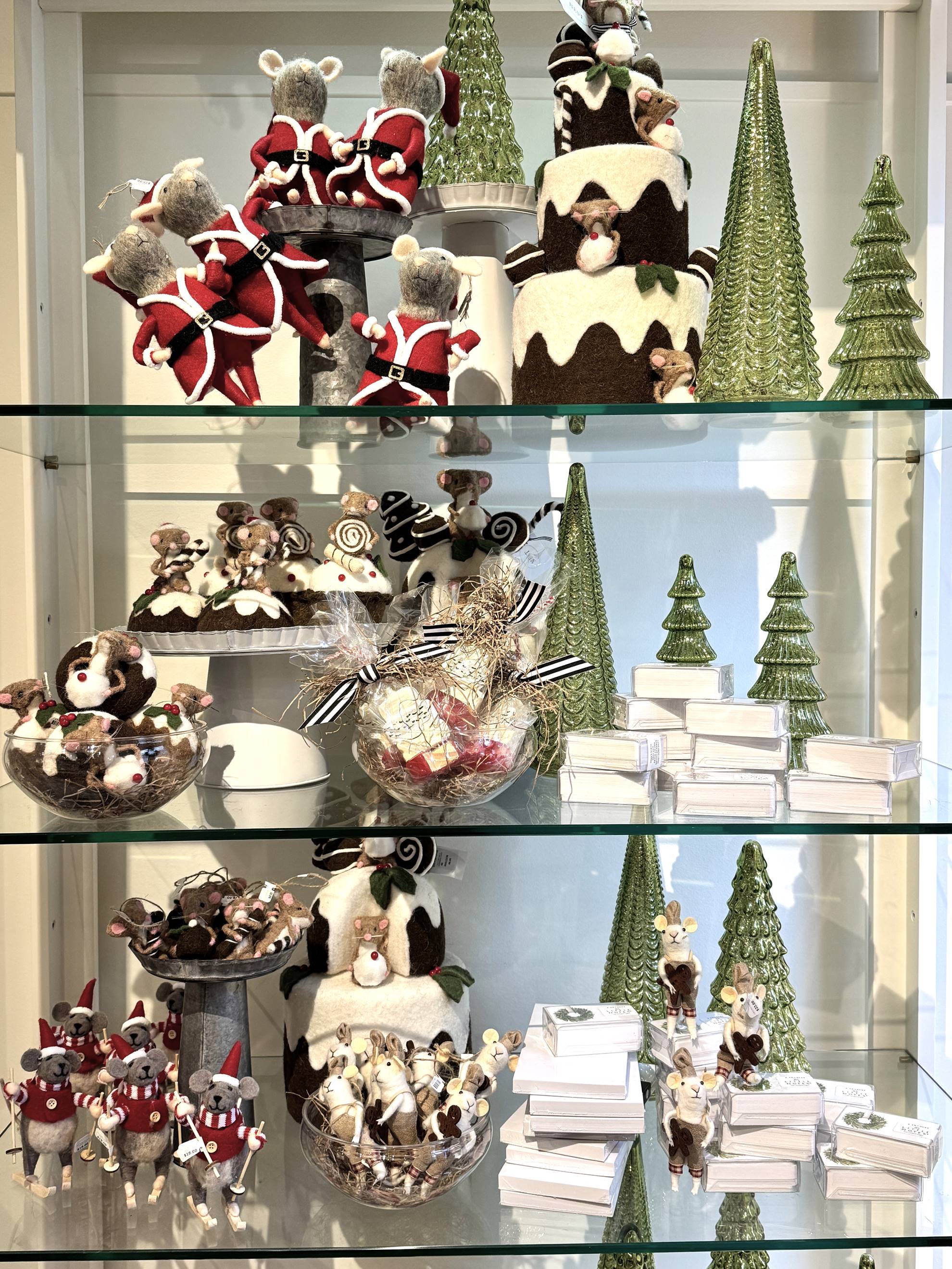 A display of Christmas decorations featuring felt mouse figurines in festive outfits, ceramic cakes, green glass Christmas trees, and stacks of white boxes arranged on three glass shelves.