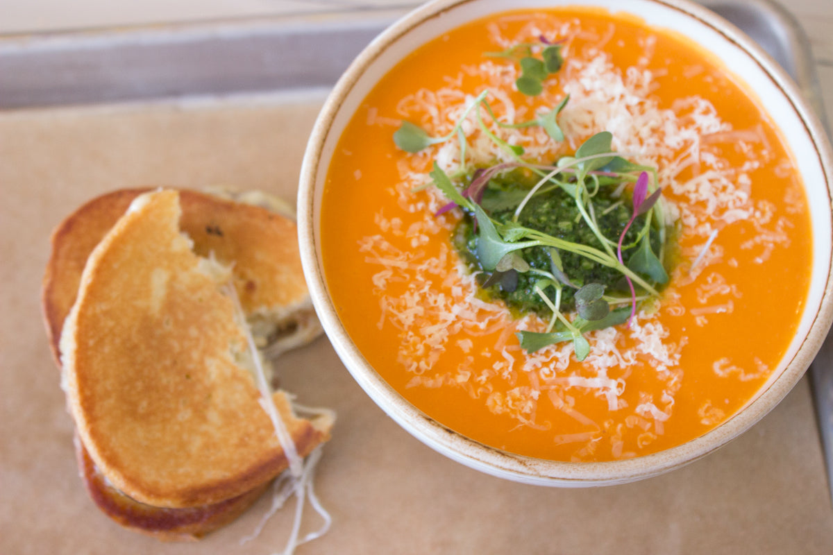A bowl of creamy tomato soup topped with shredded cheese, microgreens, and pesto, served next to a grilled cheese sandwich on a tray lined with brown paper.