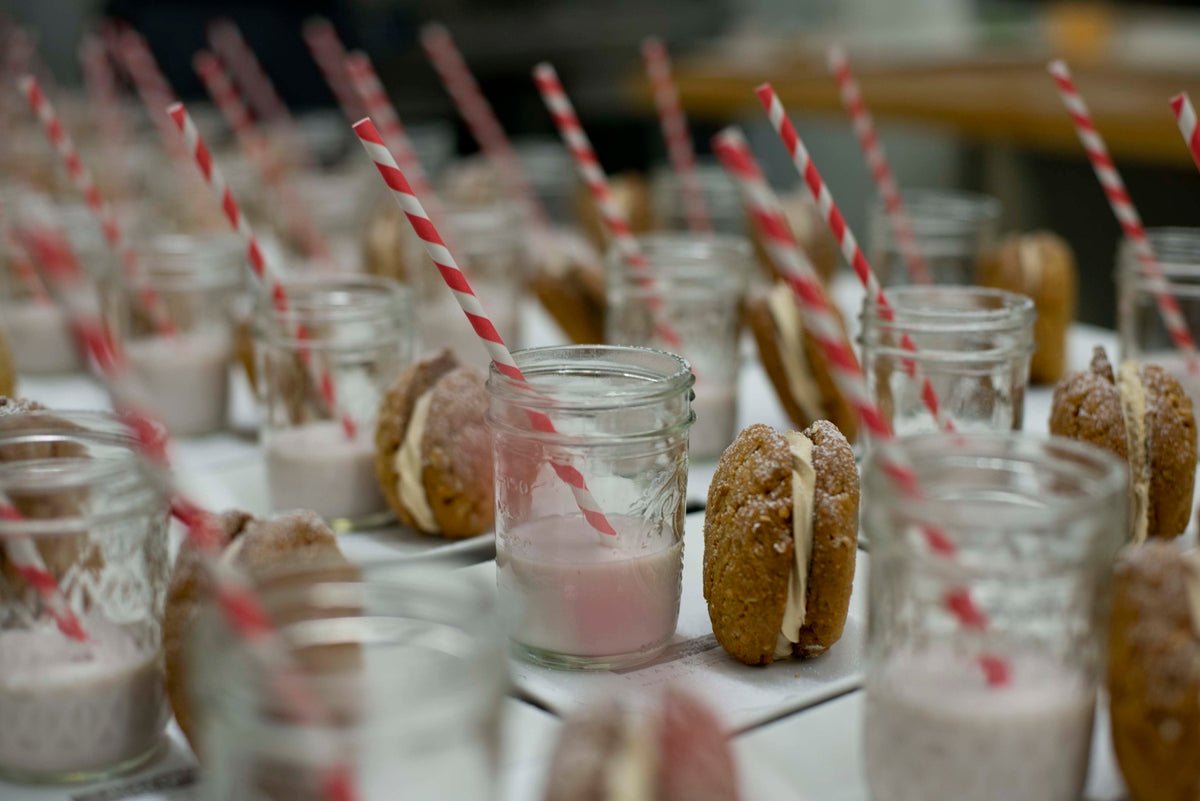 Rows of mason jars with pink milk and red-striped straws are paired with sandwich cookies, arranged neatly on a table, ready to be served.