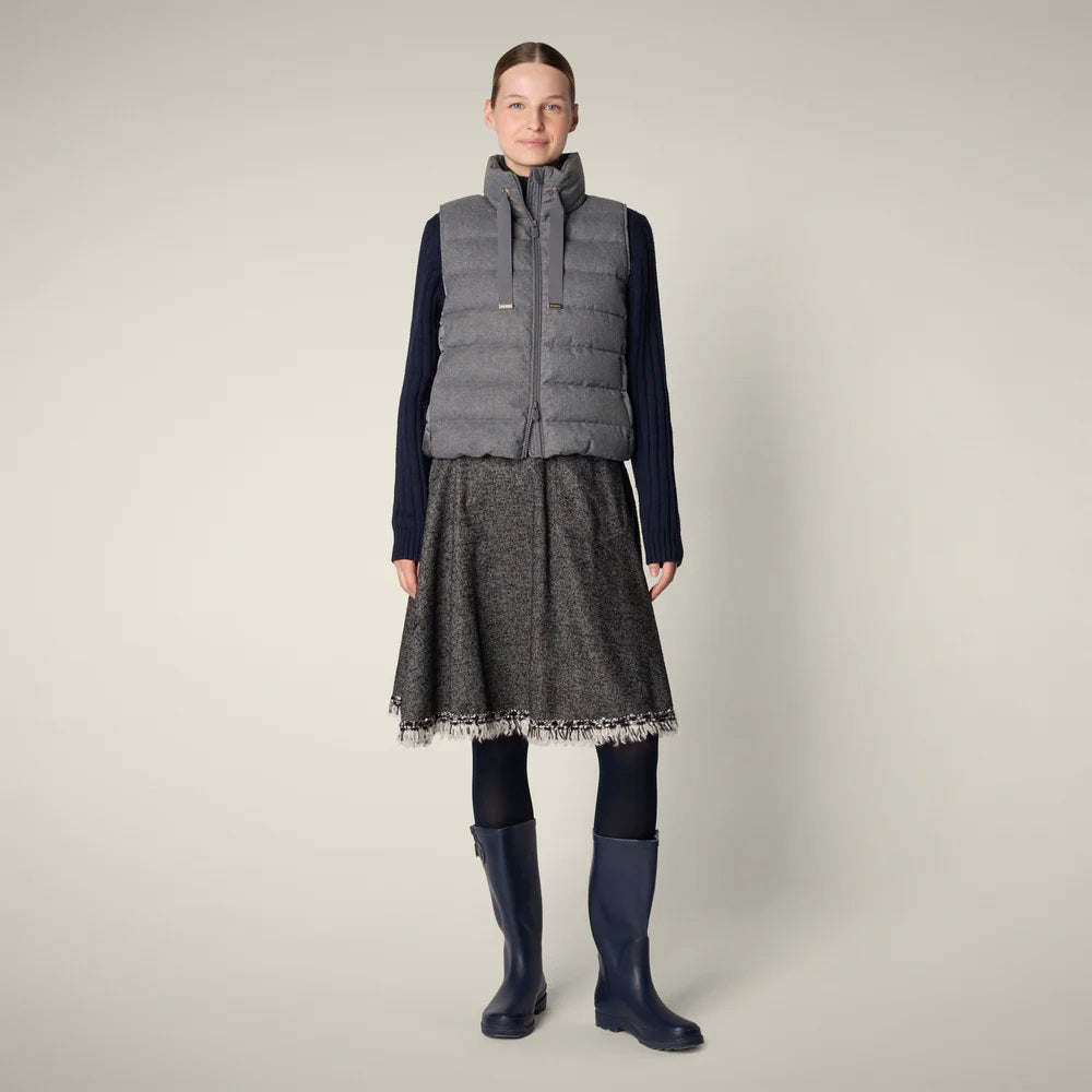A woman stands facing forward, wearing a grey puffer vest, dark long-sleeve top, grey textured skirt with fringe hem, dark tights, and navy rain boots against a plain light background.