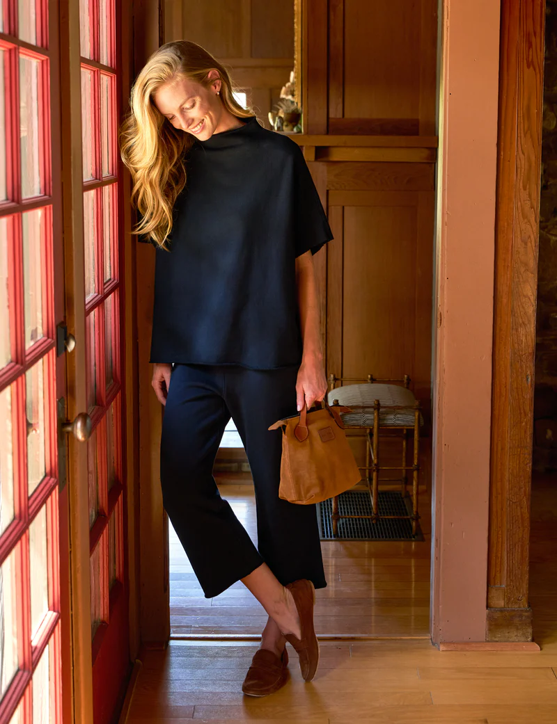 A woman with long blonde hair stands by a door, smiling and looking down. She wears a loose black top, black pants, brown shoes, and holds a brown handbag. Warm wooden interior and soft sunlight create a cozy atmosphere.