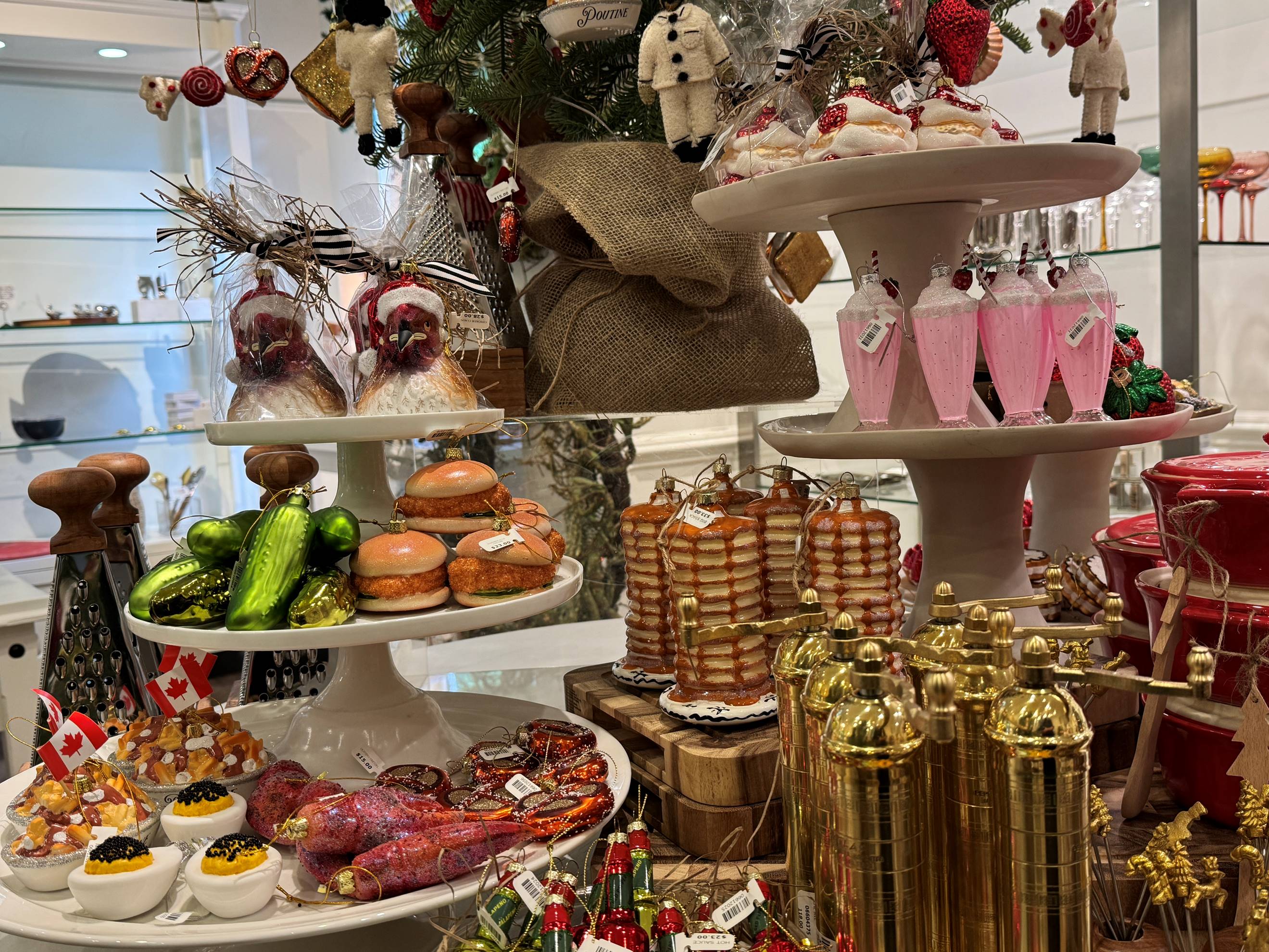 An assortment of miniature food and drink ornaments are displayed on tiered trays, including pancakes, donuts, meats, and drinks, with festive holiday decorations and small Canadian flags in the background.