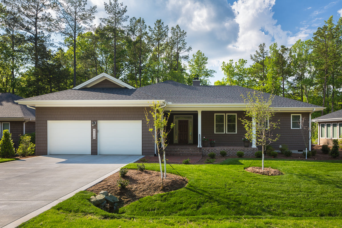 Single-story suburban house with a gray exterior, double garage, front porch, and neatly landscaped green lawn with young trees and shrubs, set against a backdrop of tall trees under a partly cloudy sky.