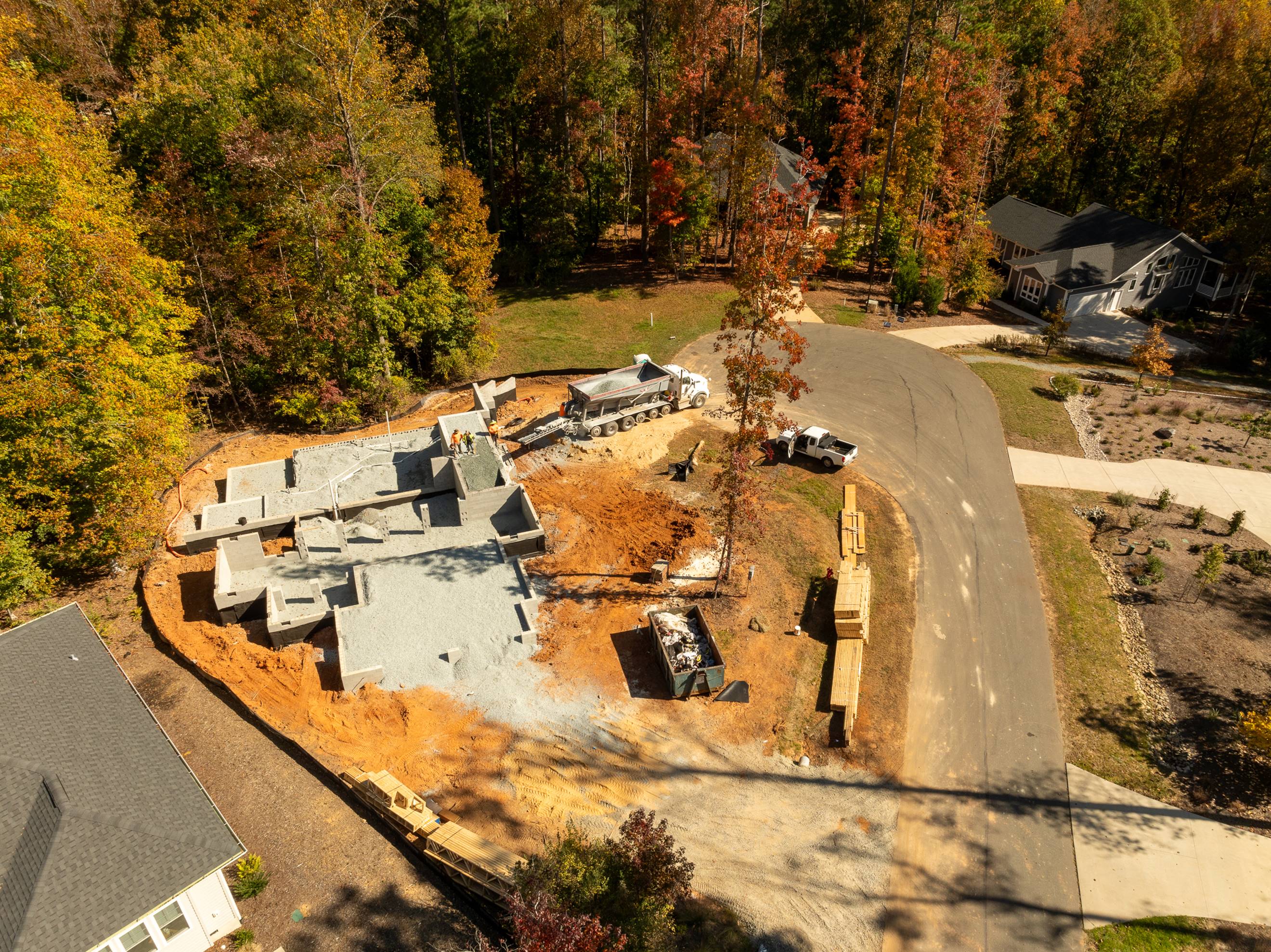 Aerial view of a house under construction in a wooded suburban neighborhood, with construction vehicles, building materials, and partially built foundation visible next to a curved road.