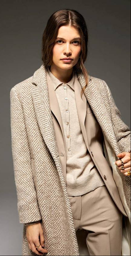 A person with long brown hair wears a beige herringbone coat over a tan suit and buttoned shirt, posing confidently with one hand in their pocket against a neutral background.