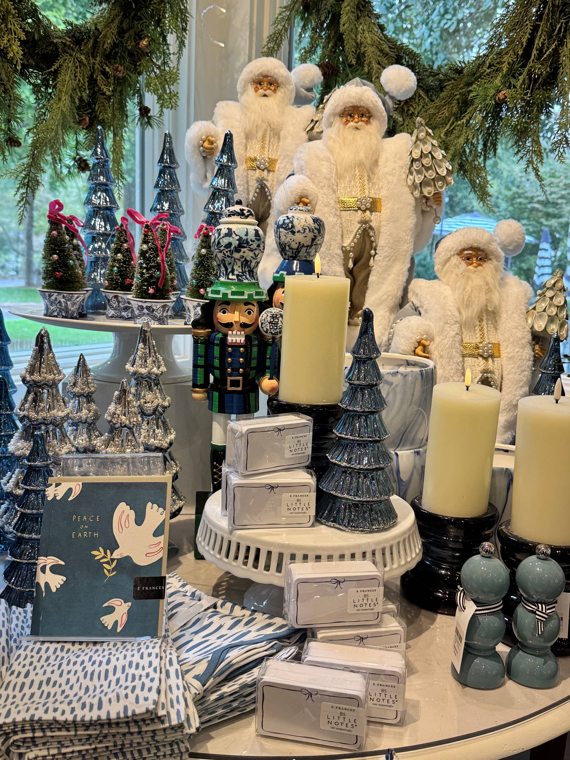 A festive display with Santa figurines, blue ceramic Christmas trees, white pillar candles, nutcracker figures, soaps, napkins, and greenery, arranged on stands in front of a window with trees outside.