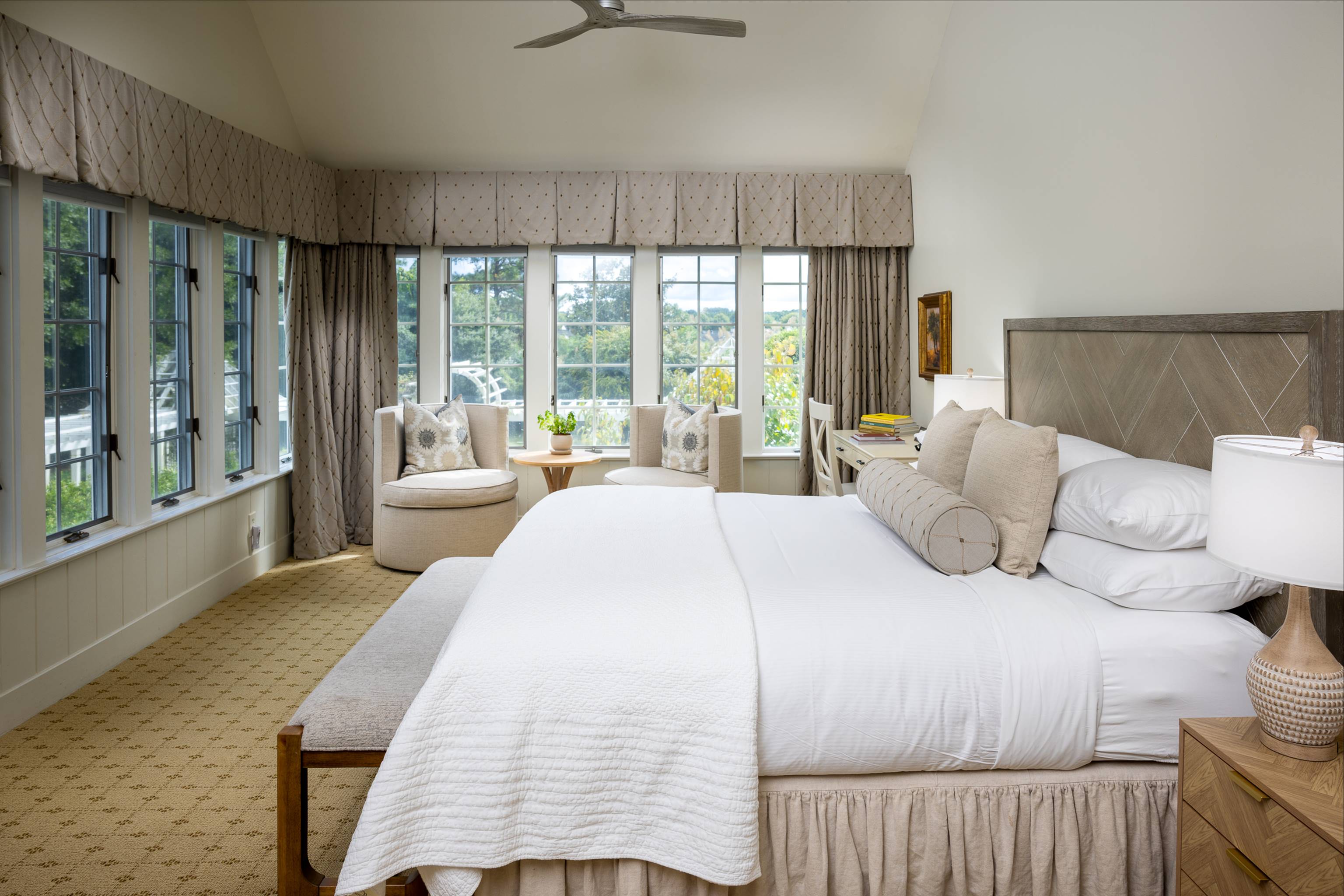 A bright, elegant bedroom with large windows, beige curtains, a king-sized bed with white bedding and neutral pillows, a bedside lamp, an armchair with a small table, and a scenic outdoor view.