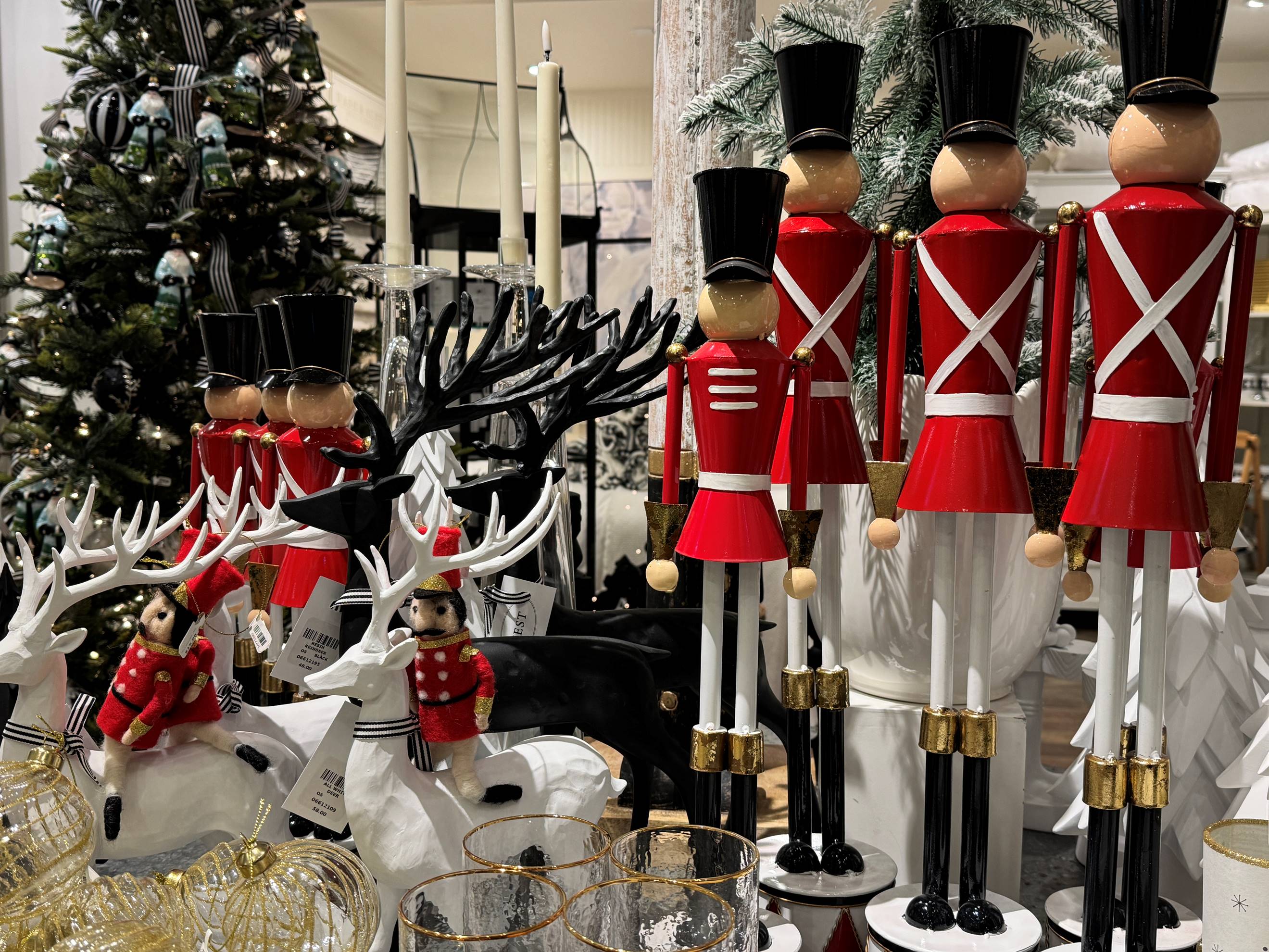A festive display with red and white toy soldiers, white and black reindeer figurines, and small dolls in red outfits, surrounded by Christmas trees and holiday decorations.