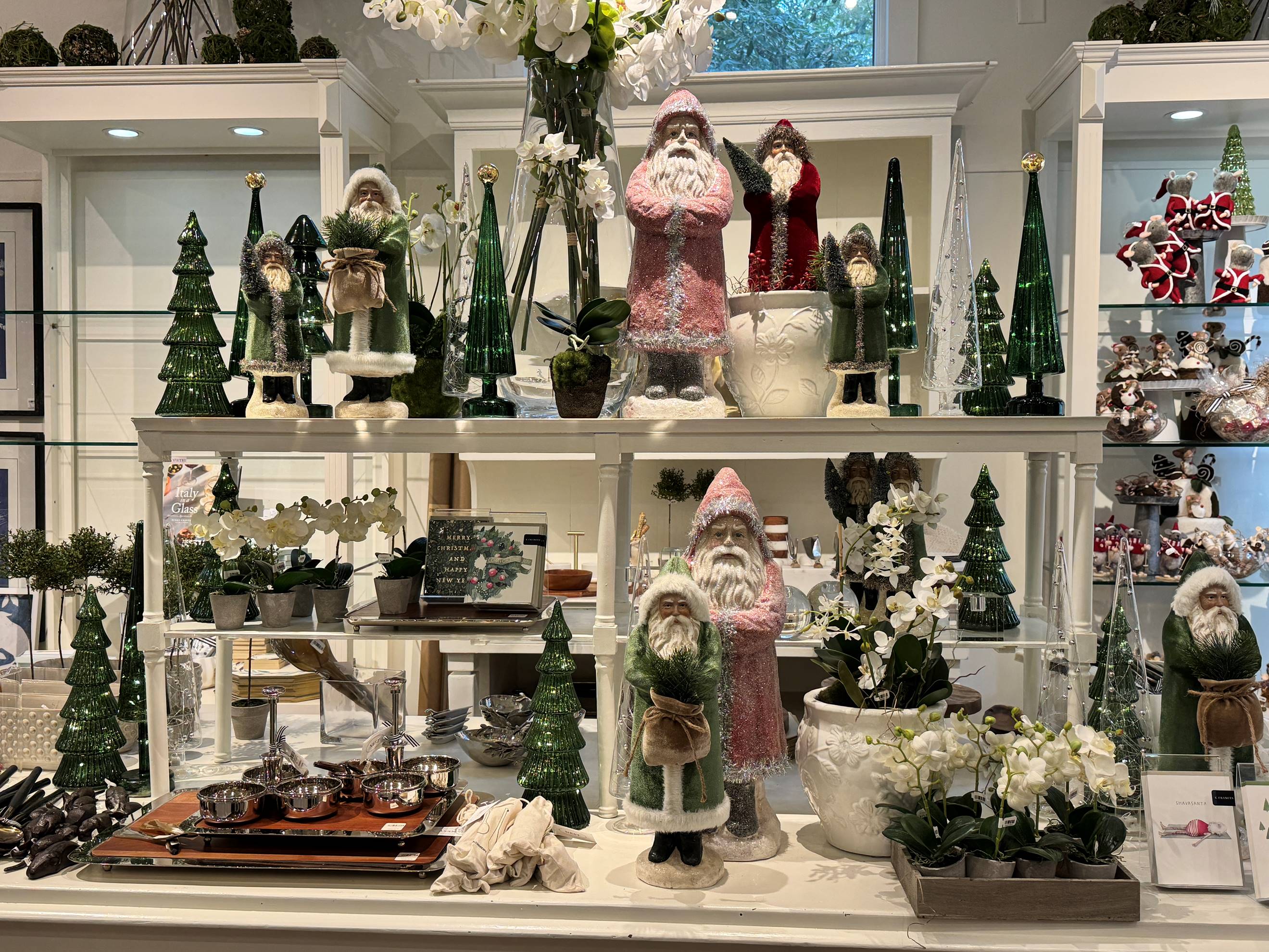 A store display features ceramic Santa Claus figures, green Christmas trees, and white floral arrangements on white shelves, with small kitchenware items and decorative plants below.