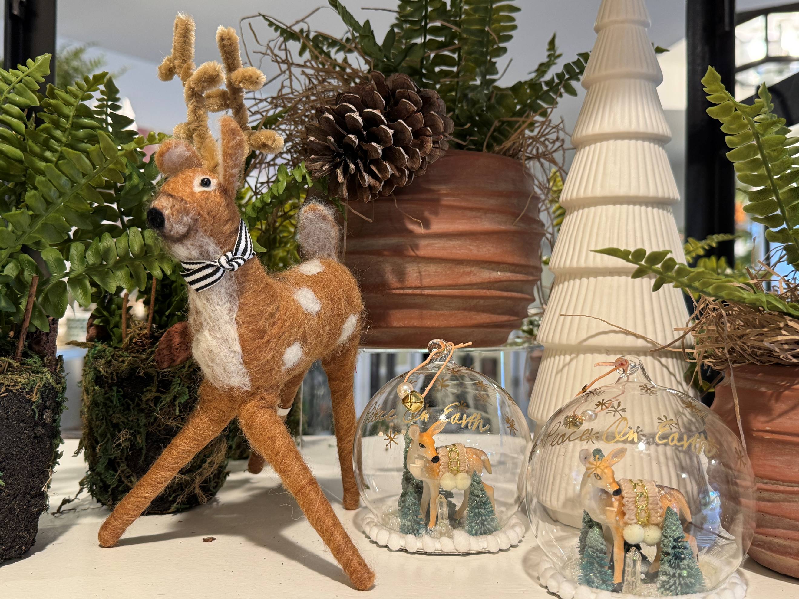A felt reindeer with a ribbon stands beside potted ferns and pinecones. Nearby are two glass ornaments containing small angel figures, with a white tree decoration in the background.