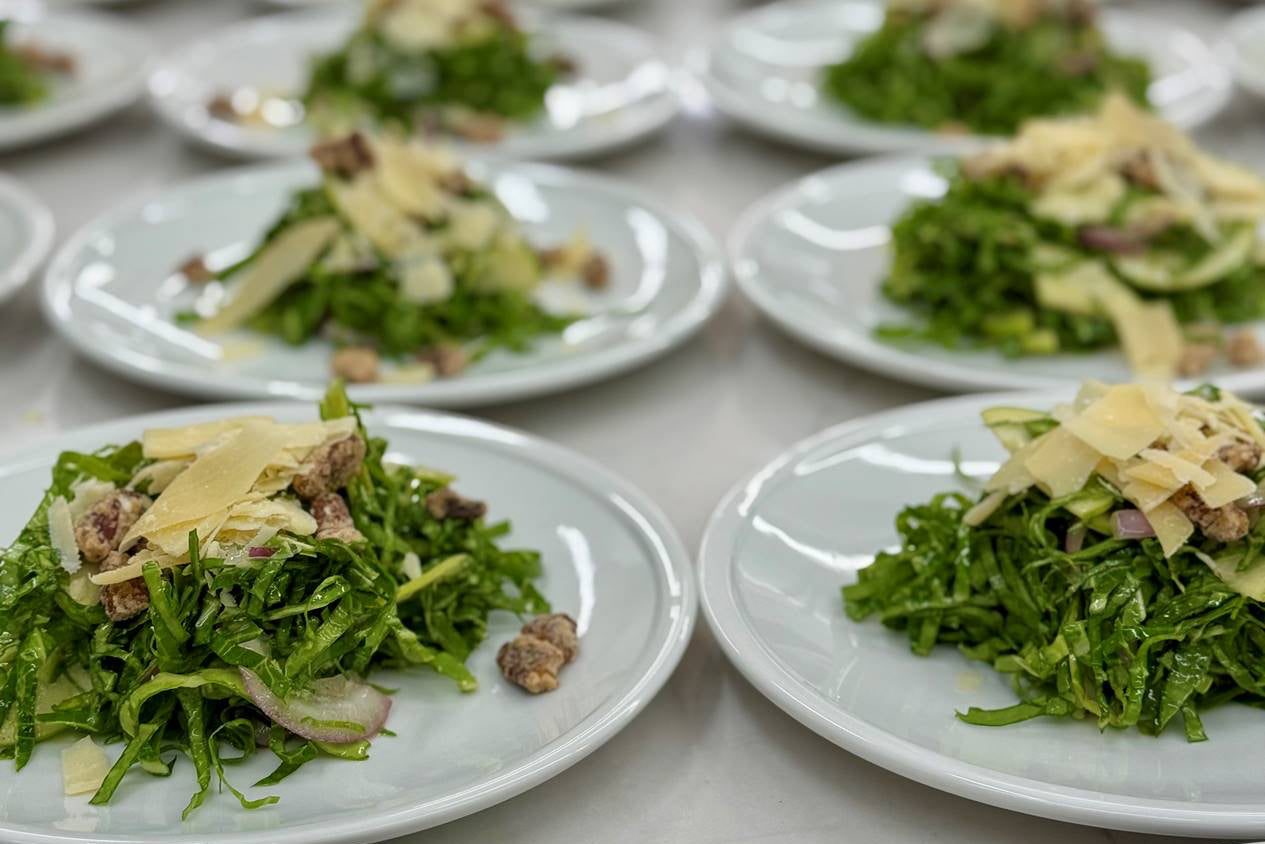 Several white plates are arranged with fresh green salads, each topped with shaved cheese, sliced red onions, and pieces of nuts, set on a light-colored surface.