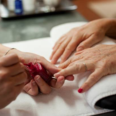 A person is getting their nails painted with red nail polish by another person. Their hands rest on a white towel, and the scene appears to be in a salon or spa setting.