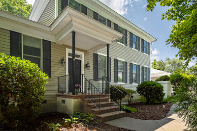 A two-story house with light-colored siding, black shutters, and a covered front porch. Brick steps with a black railing lead to the front door, surrounded by green shrubs and trees under a partly cloudy sky.
