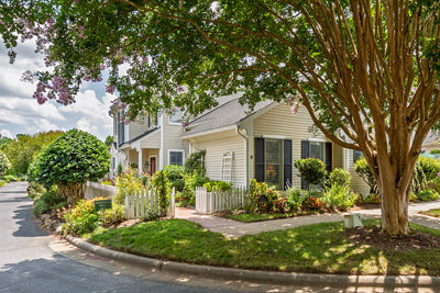 A charming house with beige siding, black shutters, and a white picket fence is surrounded by lush landscaping, flowering trees, and greenery along a quiet suburban street on a sunny day.