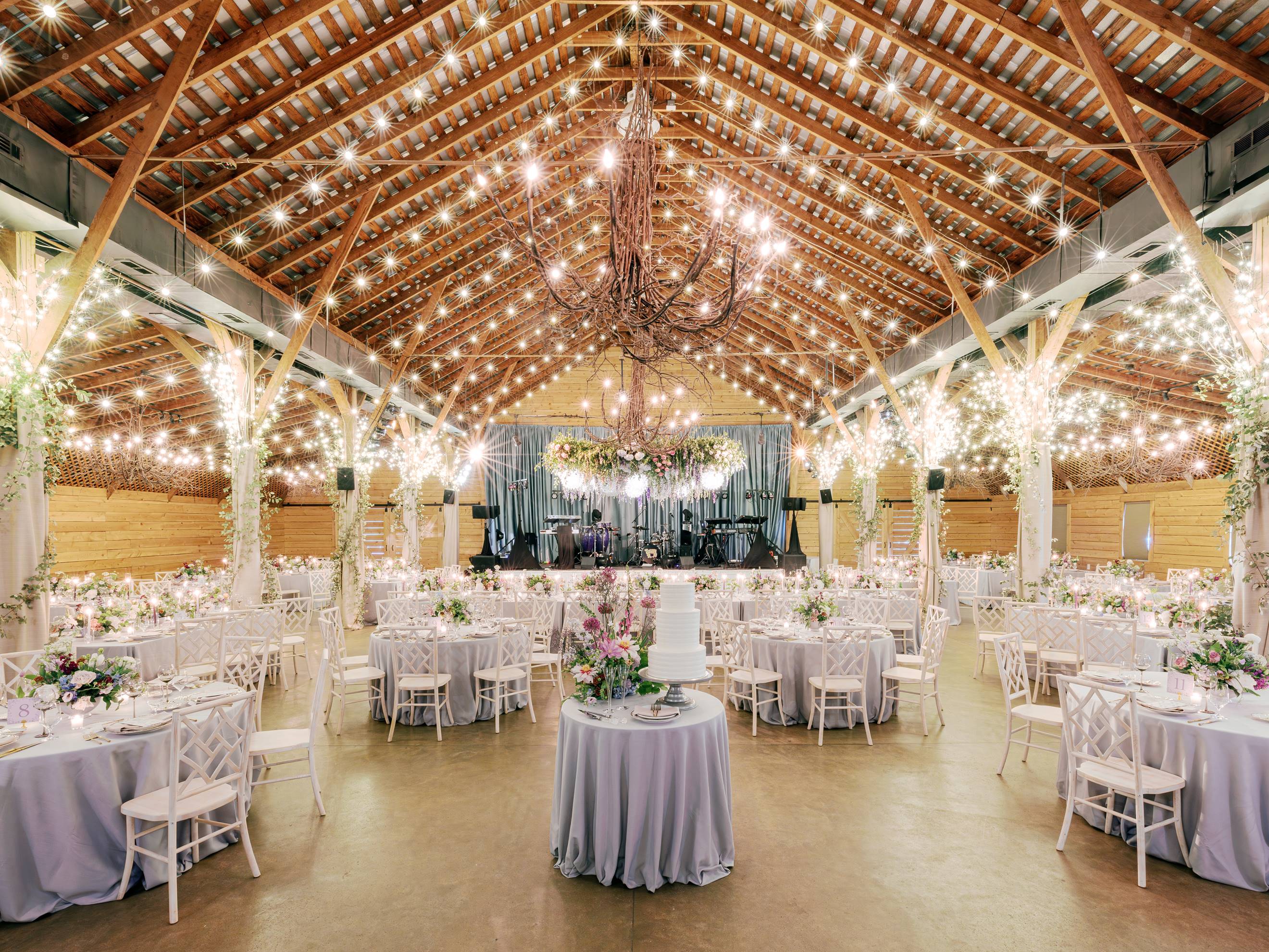 A spacious barn venue decorated for an event, featuring round tables with white chairs, floral centerpieces, a large chandelier, string lights, and a multi-tiered cake on a round table in the center.