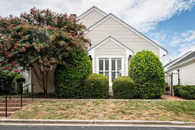 Single-story beige house with white trim, large front bushes, a flowering tree on the left, and a manicured lawn bordered by a sidewalk under a partly cloudy sky.