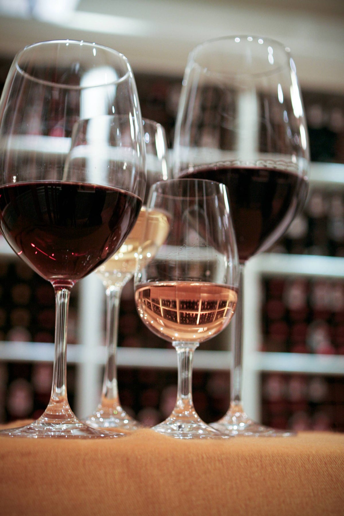 Four wine glasses containing red, white, and rosé wines are lined up on a brown surface, with wine racks blurred in the background.