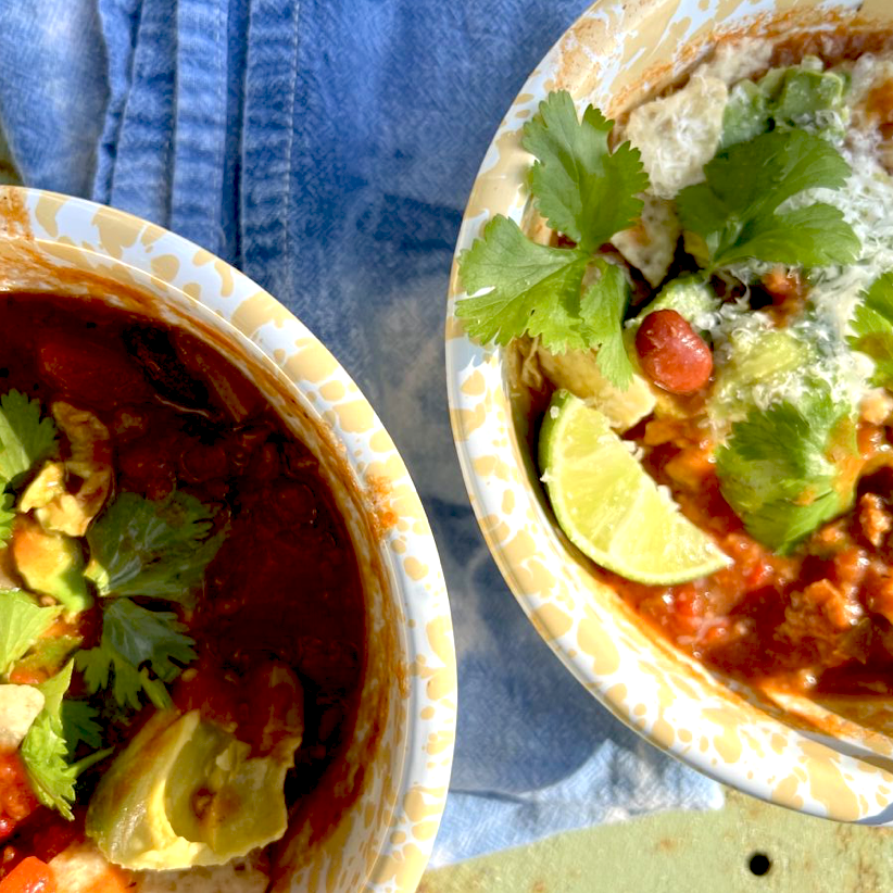 Two bowls of chili topped with avocado, lime wedges, and fresh cilantro, sitting on a blue cloth. The bowls contain a red tomato-based stew with beans and other garnishes.