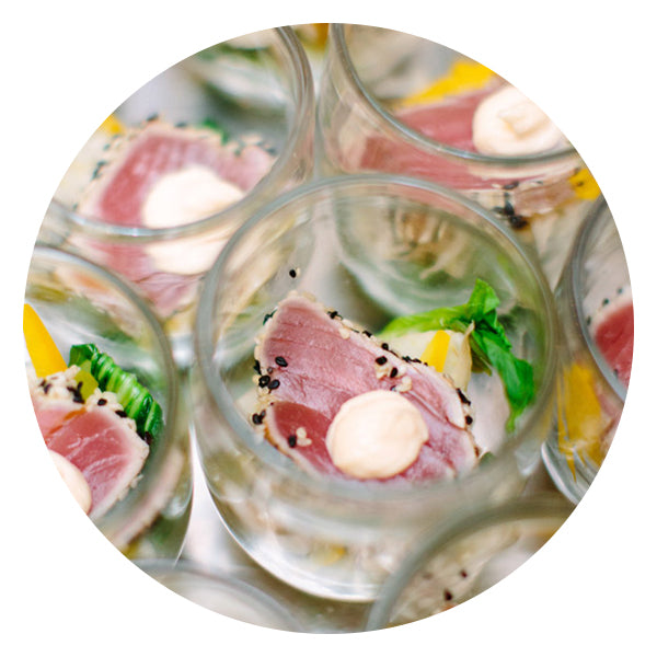 A close-up of small glass cups containing seared tuna with a dollop of sauce, garnished with sesame seeds, greens, and thin yellow pepper slices.