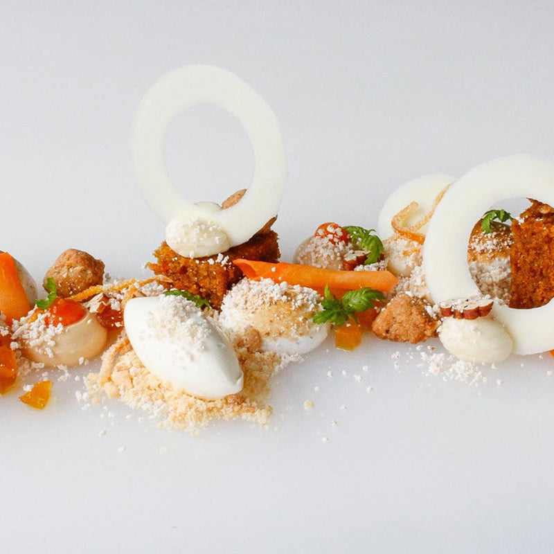 A modern dessert artfully plated features white chocolate rings, dollops of cream, crumbled cookie, cake pieces, orange segments, caramel shards, and green herb garnishes on a white surface.