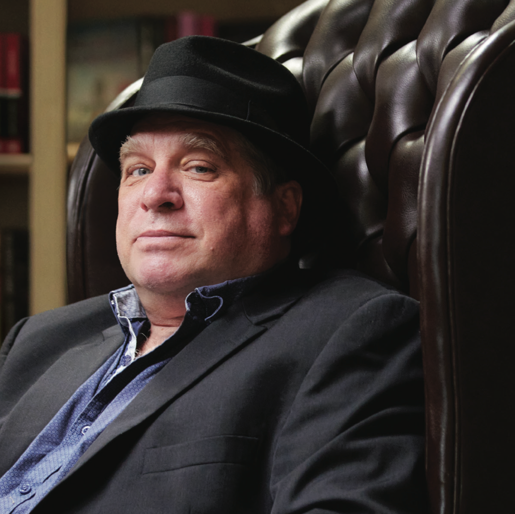 A middle-aged man in a black fedora and dark blazer sits on a brown leather armchair, looking at the camera with a neutral expression. Bookshelves are visible in the background.