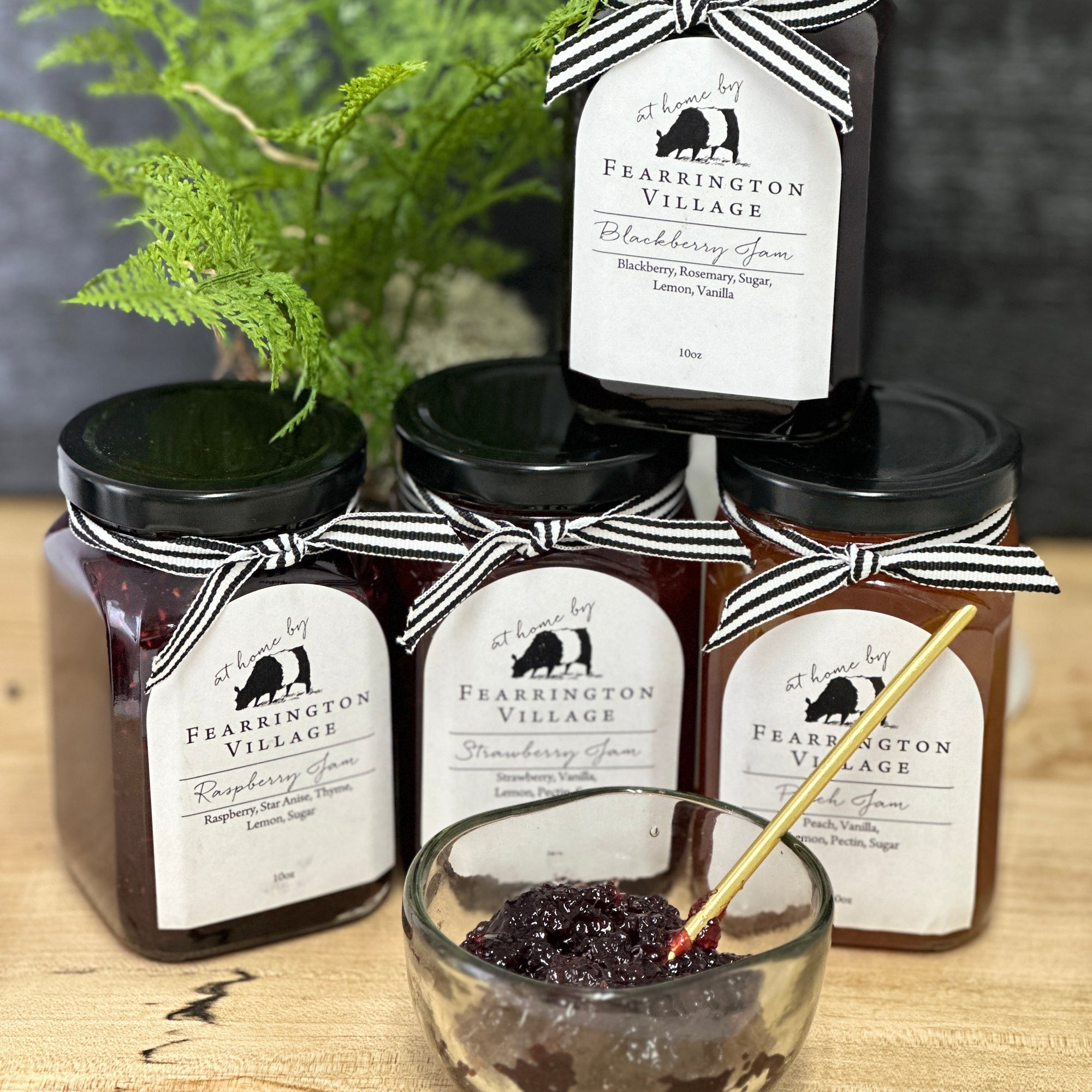 Four jars of Fearrington Village preserves with striped ribbon bows are arranged with a small glass bowl of jam and a gold spoon in front. A green fern is in the background, adding a fresh touch.