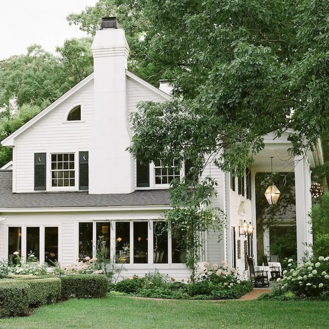A charming white house with tall windows, black shutters, and a large chimney is surrounded by green bushes, grass, and trees. A covered porch with hanging lights and outdoor seating is visible on the right.