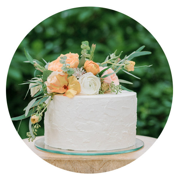 A round white frosted cake decorated with orange and white flowers and greenery sits on a glass plate, with a blurred green background.