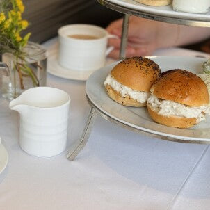 A close-up of two sandwiches filled with a creamy spread on a tiered tray, next to a white coffee cup, a small white pitcher, and a vase with yellow flowers on a white tablecloth.