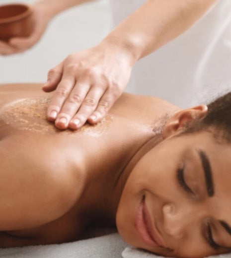 A woman lies on her stomach, smiling, while receiving a back massage with a scrub applied to her skin by a massage therapist’s hand.