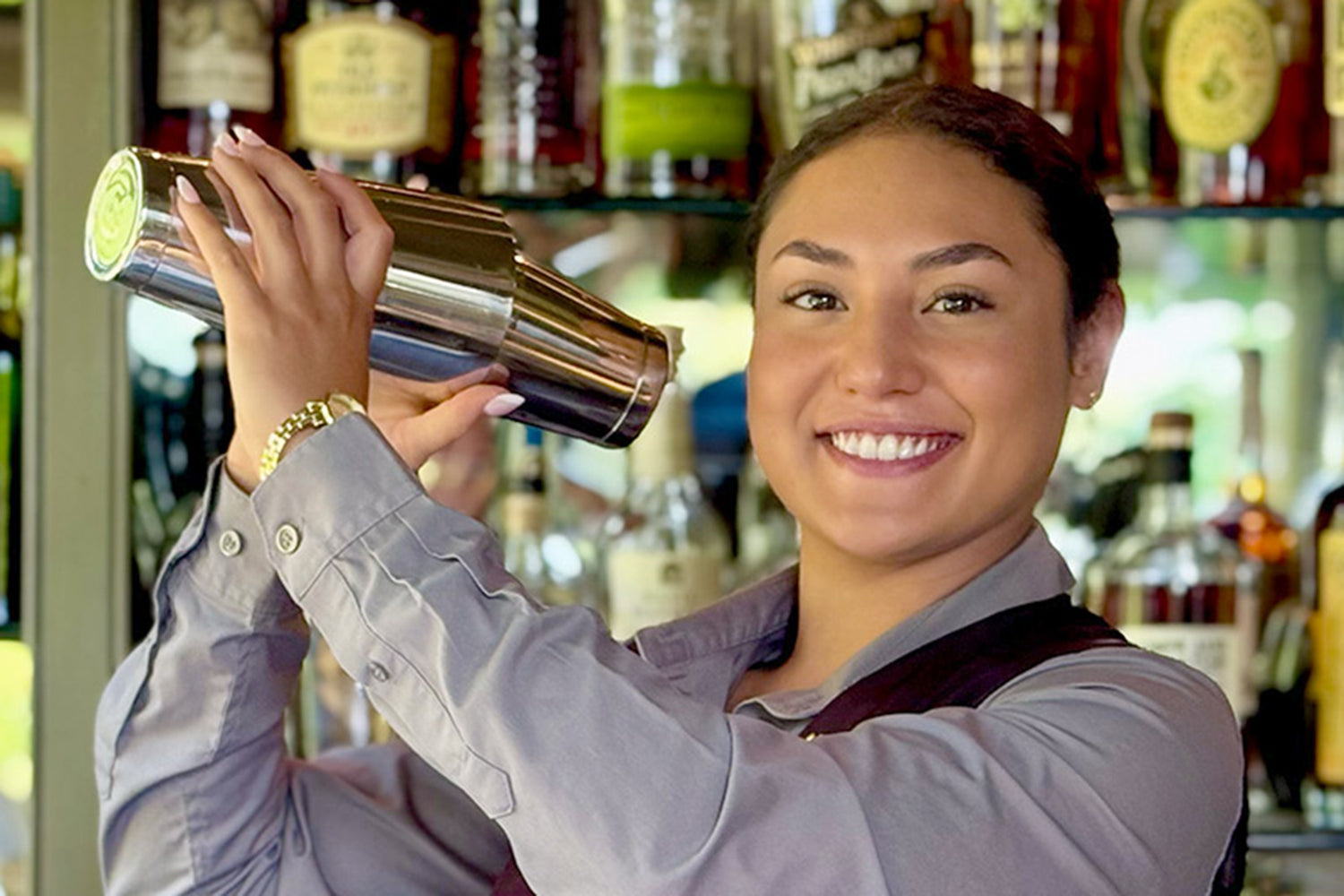 A smiling bartender in a gray shirt shakes a cocktail shaker behind a bar lined with various bottles of liquor.