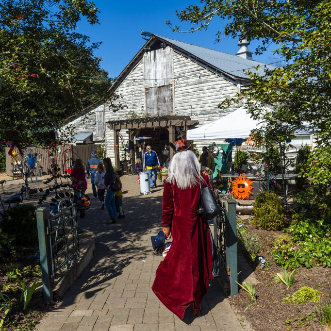 A person in a long red coat walks toward a rustic barn where people are gathered outside, surrounded by greenery and various outdoor decorations on a sunny day.