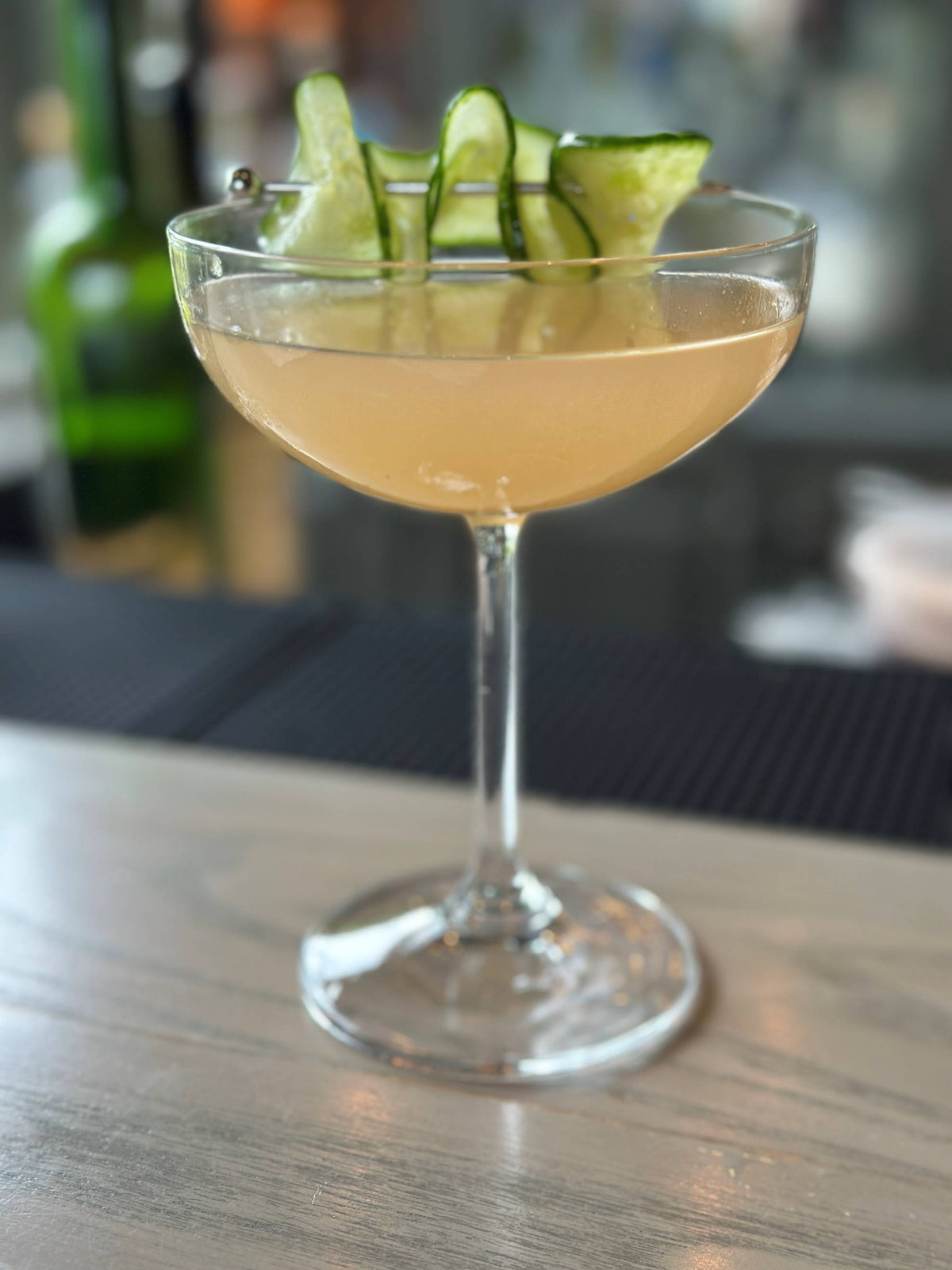 A pale yellow cocktail in a coupe glass, garnished with thinly sliced cucumber ribbons, sits on a light wooden bar top with a blurred background.