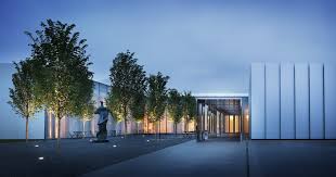Modern building exterior at dusk with tall white walls, illuminated windows, and a row of trees along a walkway. Outdoor lights highlight a sculpture near the entrance, creating a serene atmosphere.