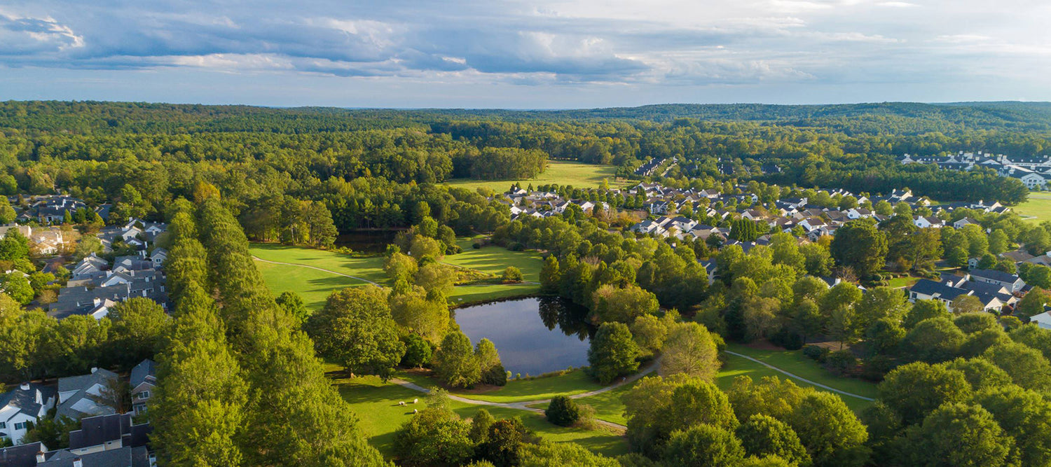 Aerial view of a suburban neighborhood surrounded by dense green trees and a small pond in the center, with houses scattered throughout the landscape under a partly cloudy sky.