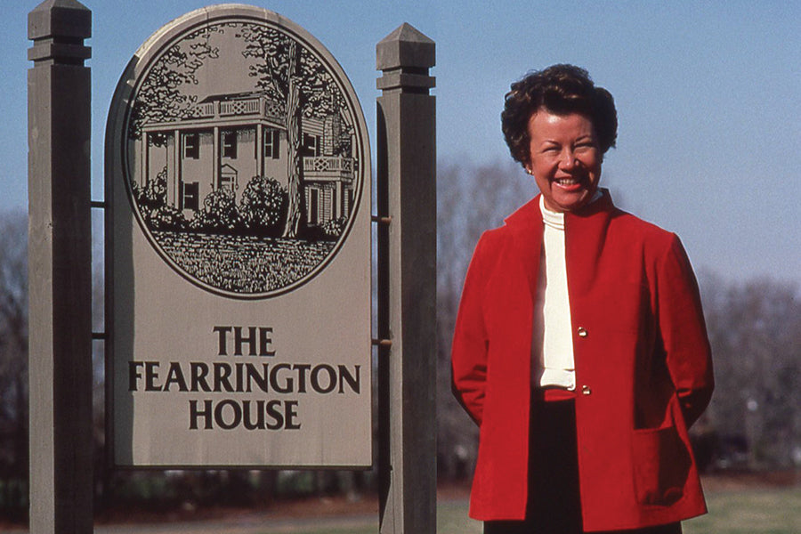 A woman in a red jacket and white blouse stands smiling next to a large sign that reads 