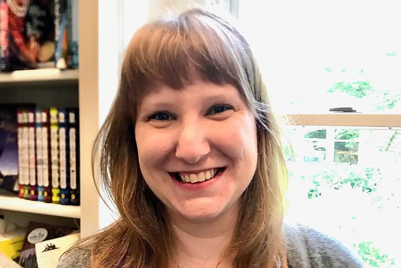 A person with light brown hair and bangs smiles in front of a window. Books are visible on a shelf to the left, and daylight comes through the window in the background.