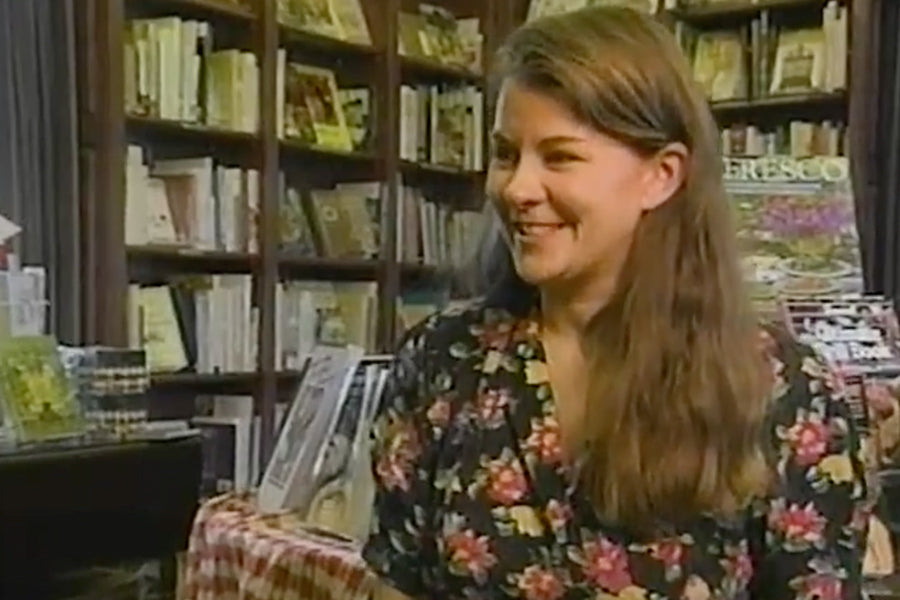 A woman with long brown hair wearing a floral dress smiles while sitting in a bookstore, with shelves of books and magazines visible in the background.