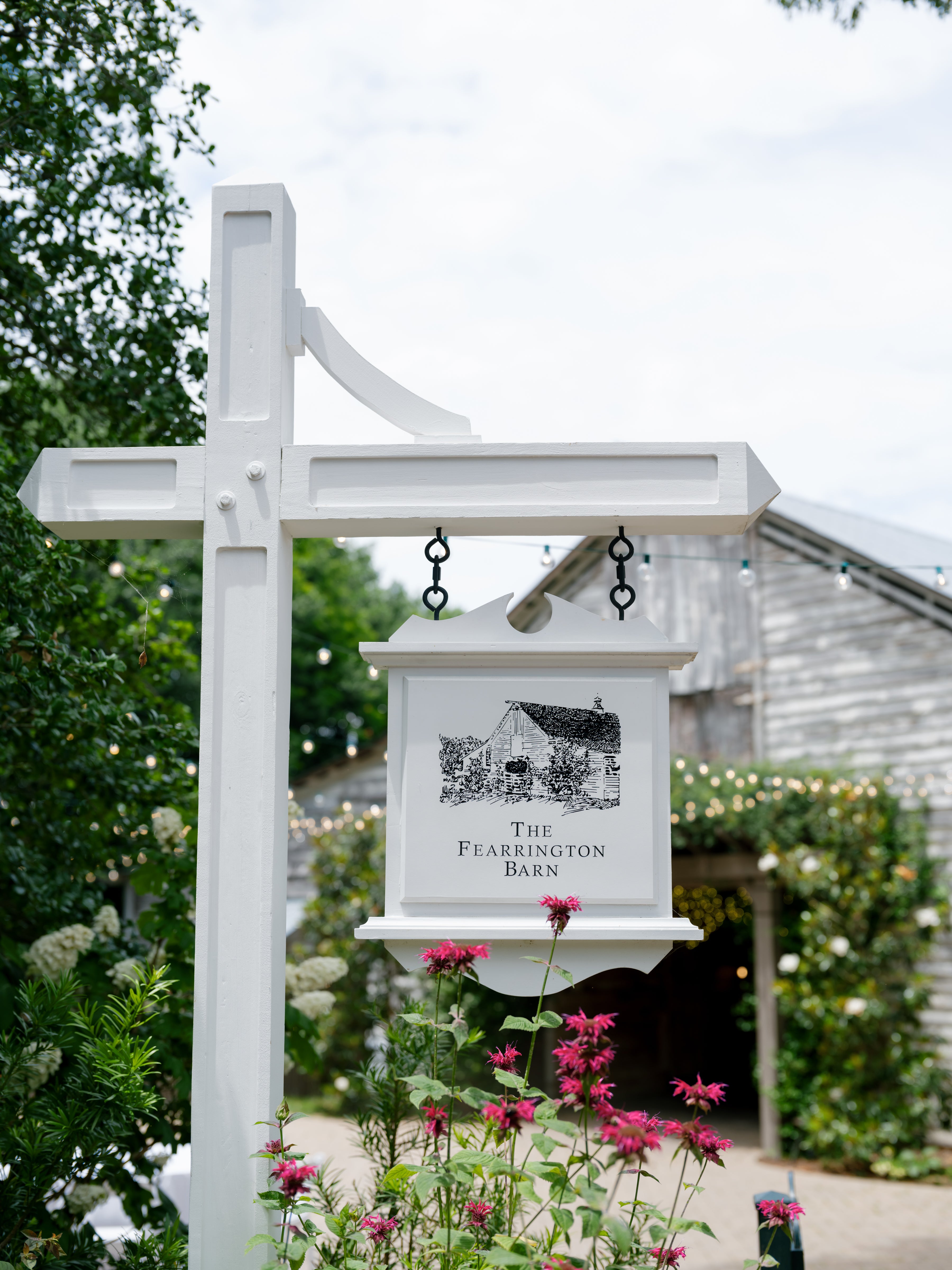 A white wooden signpost with a hanging sign reading “The Fearrington Barn” stands by pink flowers in front of a rustic barn covered in greenery and string lights.
