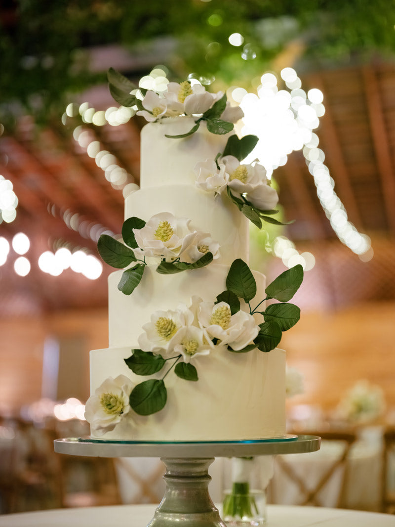 A four-tiered white wedding cake decorated with realistic white flowers and green leaves, displayed on a silver stand with blurred lights and greenery in the background.