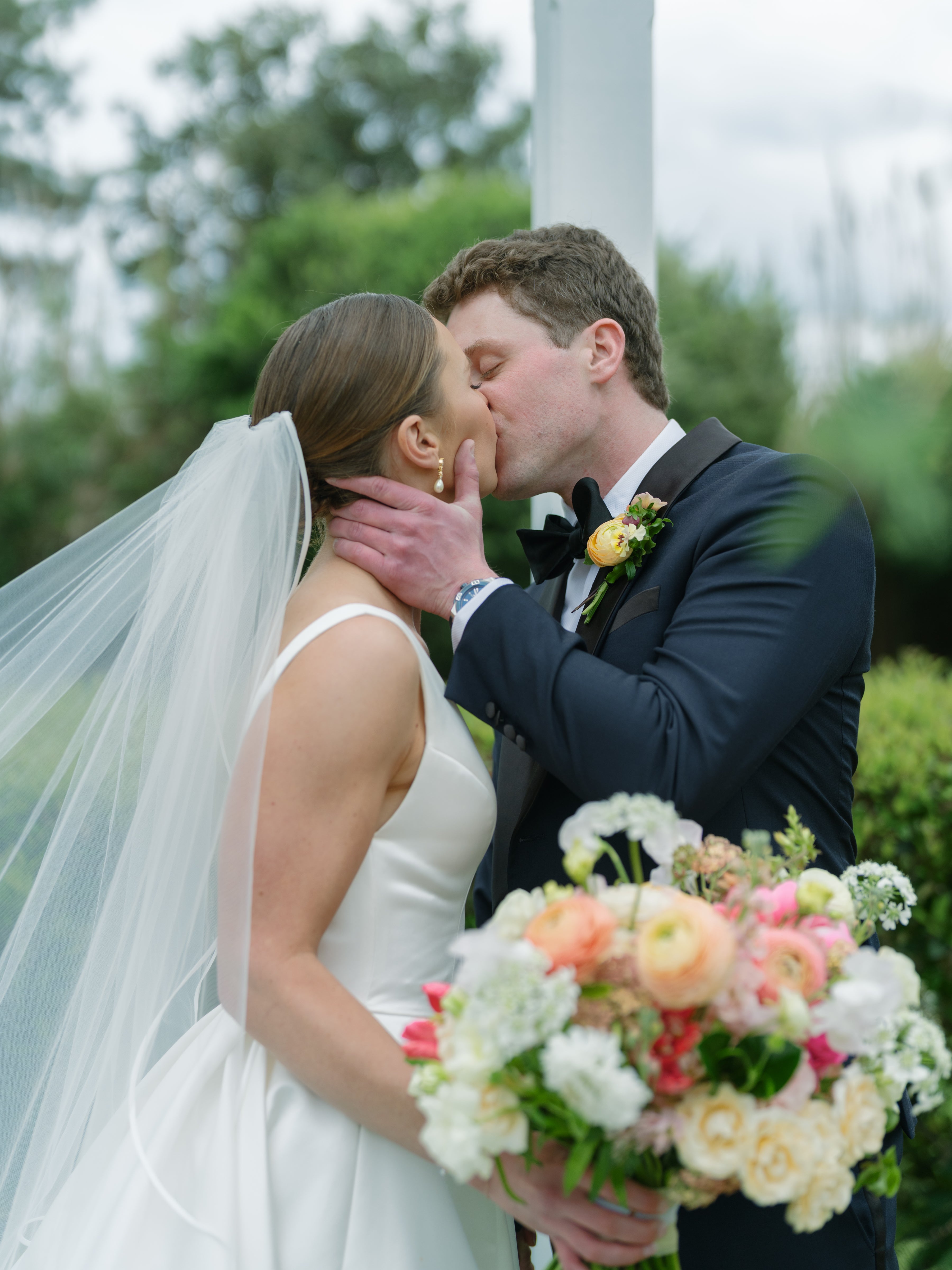A bride and groom share a kiss outdoors. The bride wears a white dress and veil, holding a colorful bouquet. The groom is in a dark suit with a boutonniere. Greenery and trees are visible in the background.