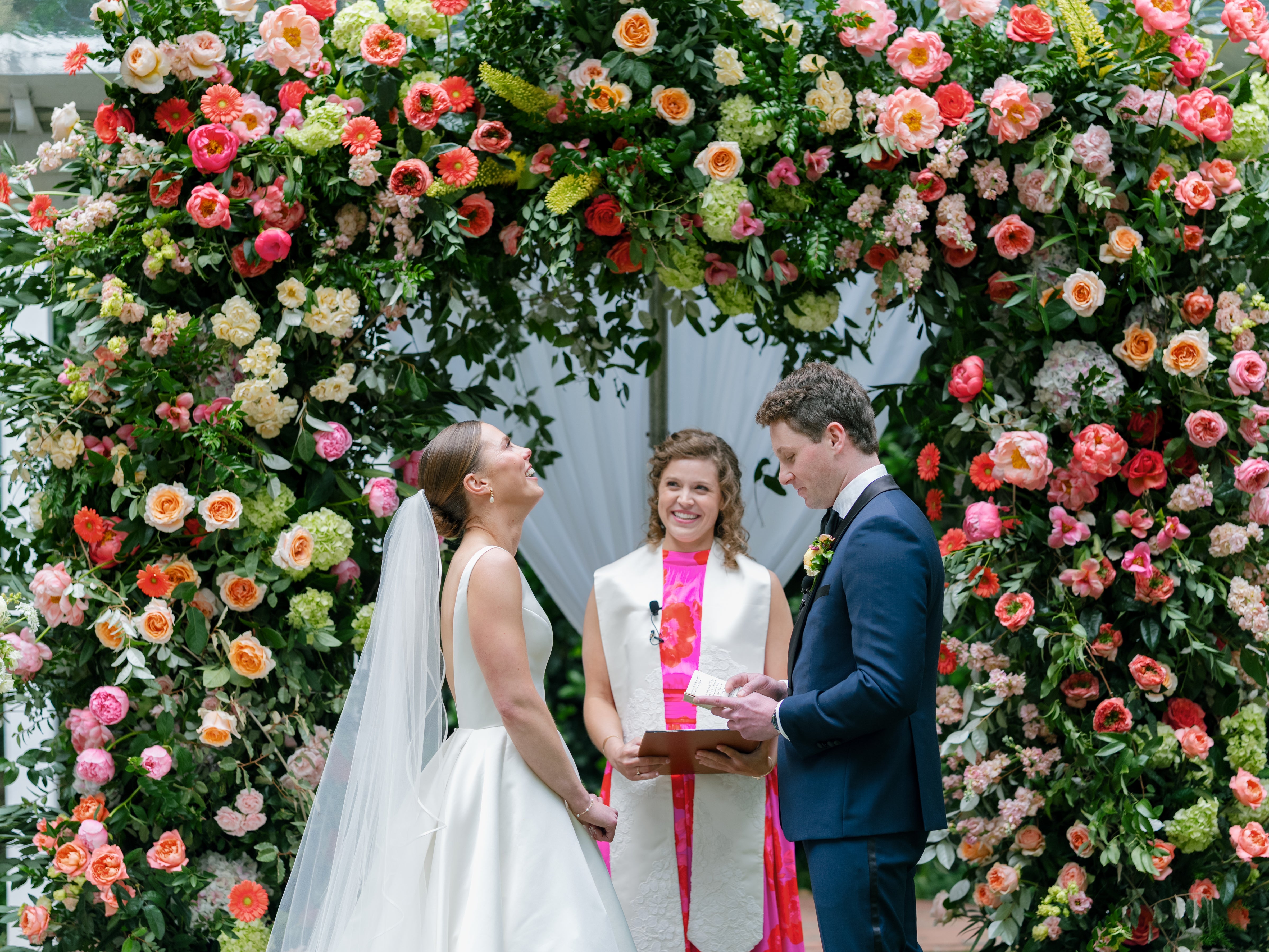 A bride and groom stand under a vibrant floral arch, smiling with an officiant between them during an outdoor wedding ceremony.