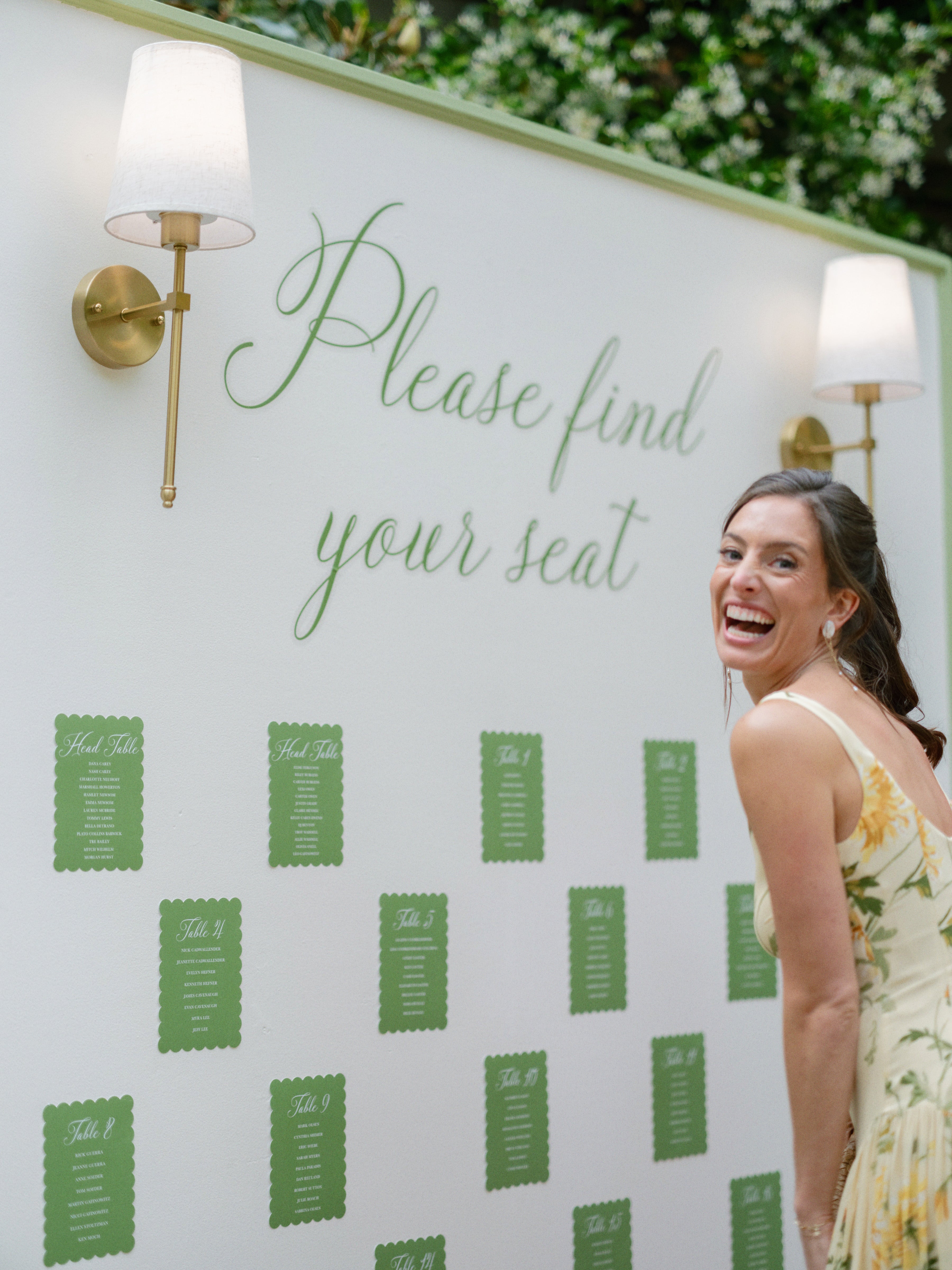 A smiling woman in a yellow floral dress stands beside a seating chart board that reads “Please find your seat,” with lists of names displayed under wall sconces.