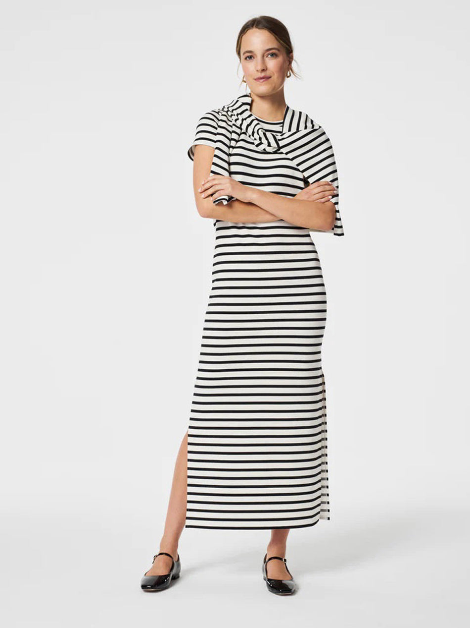 A woman stands with arms crossed, wearing a black and white striped maxi dress with short sleeves and a side slit. She has her hair tied back and is wearing black flat shoes, posing against a plain white background.