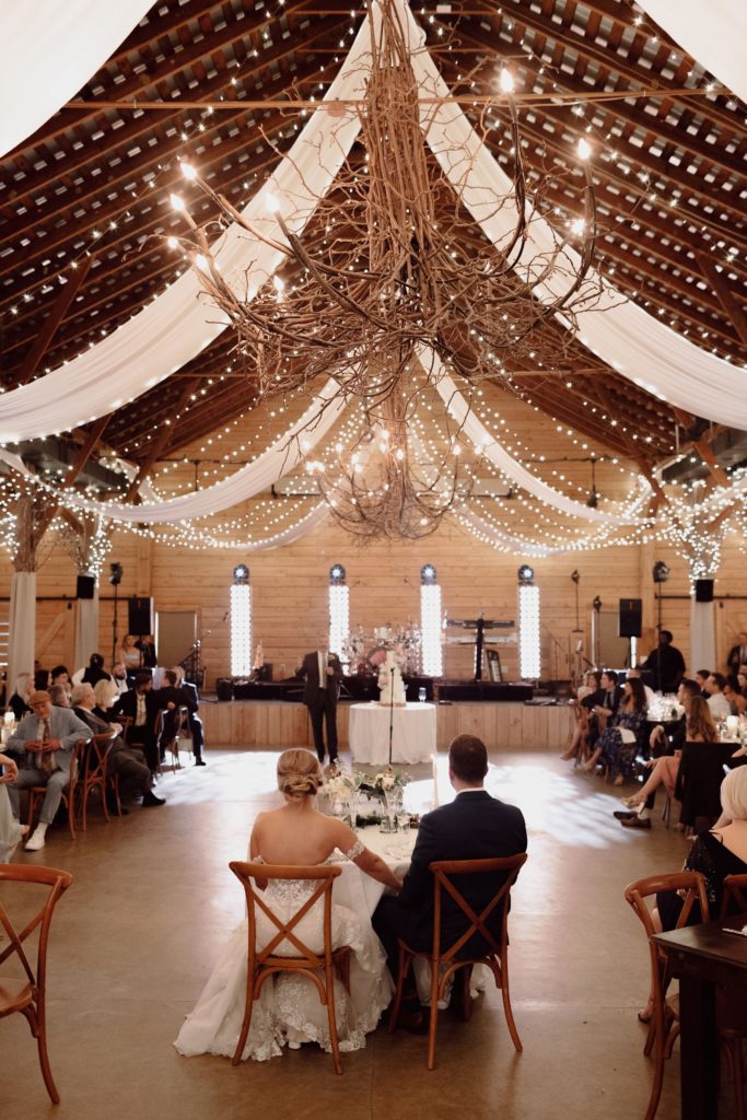 A bride and groom sit hand-in-hand facing their wedding cake in a rustic barn decorated with fairy lights, draped white fabric, and a large twig chandelier, surrounded by seated guests.