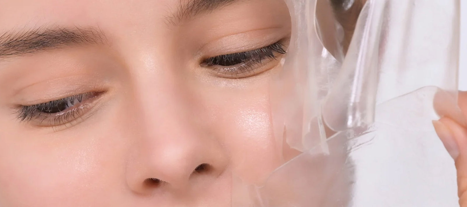 Close-up of a person gently peeling a transparent facial mask from their smooth, clear skin, focusing on the eye and cheek area. The mask is partially lifted, revealing a fresh, glowing complexion underneath.